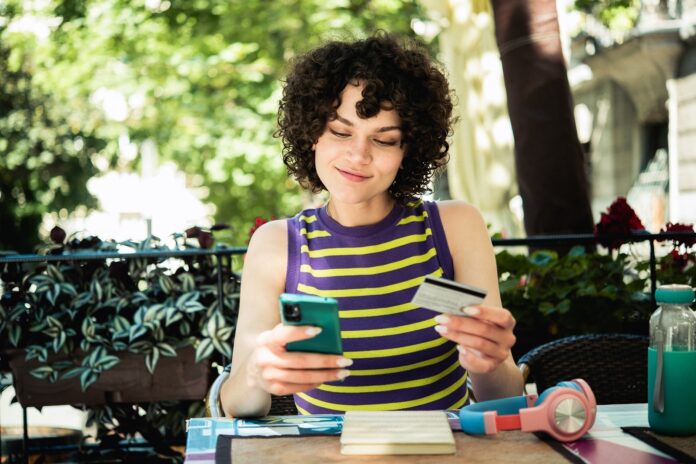 Young-college-student-sitting-outdoors-holding-credit-card-while-using-smart-phone_MixMedia.jpg